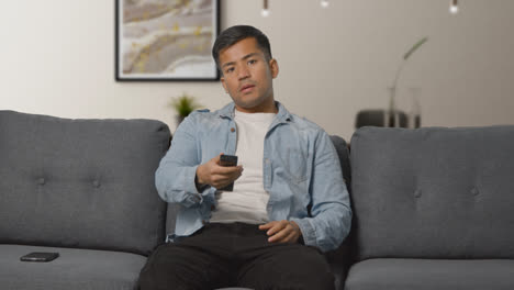Young-Man-Sitting-On-Sofa-At-Home-With-Remote-Control-Flicking-Through-TV-Channels